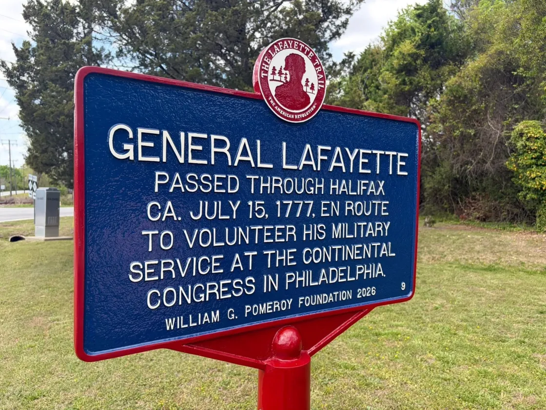 A blue historical marker outlined in red with white lettering titled “General Lafayette” stands on a lawn in front of trees commemorating Lafayette's Visit this city.