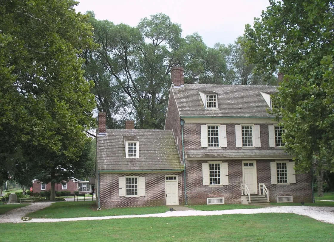 A brick colonial‑style house with cream shutters and multiple chimneys stands amid green grass and tall trees.