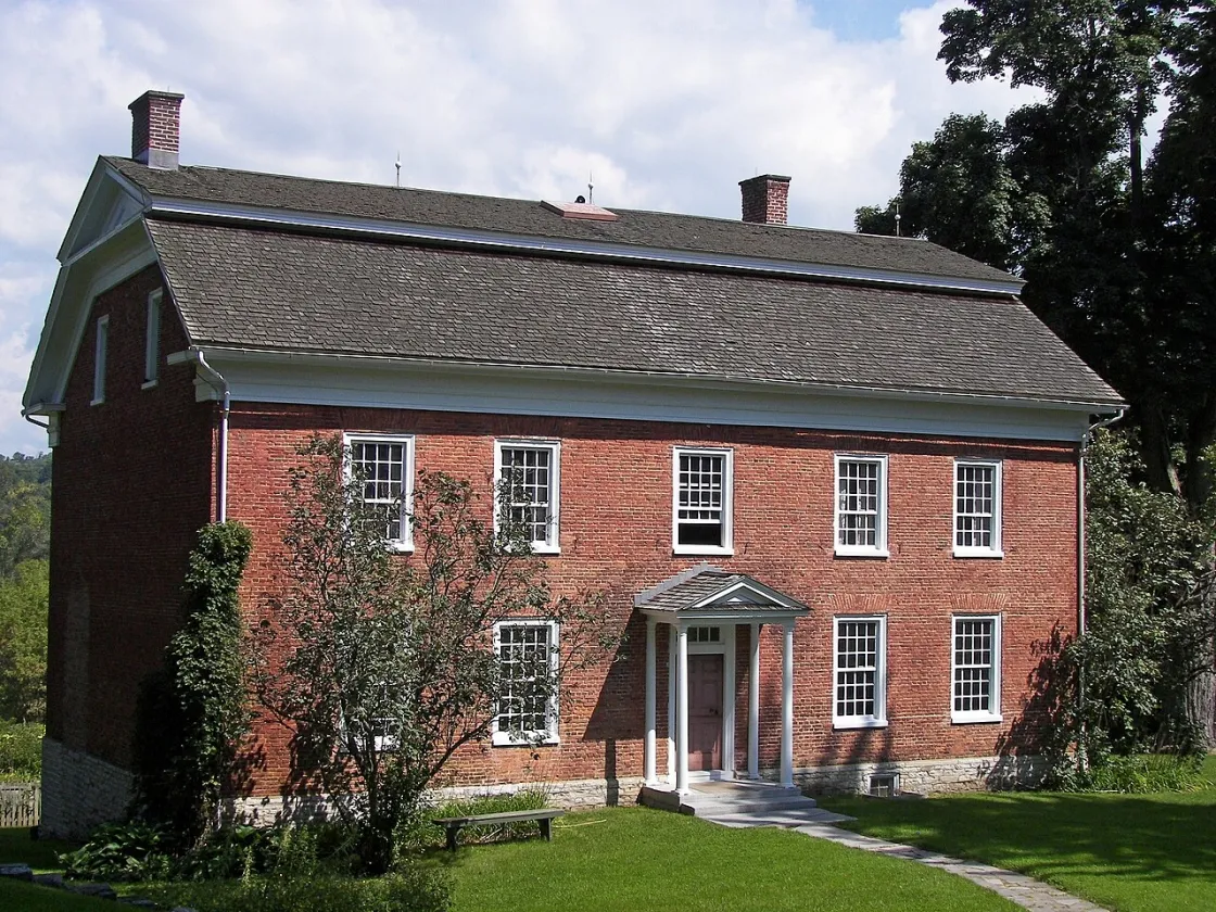 Red brick 2-story colonial home with a gambrel roof, two chimneys, and 9 windows trimmed in white.and 