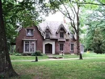 A large brick Gothic‑style house with steep gables and tall narrow windows stands among mature trees on a grassy lawn.