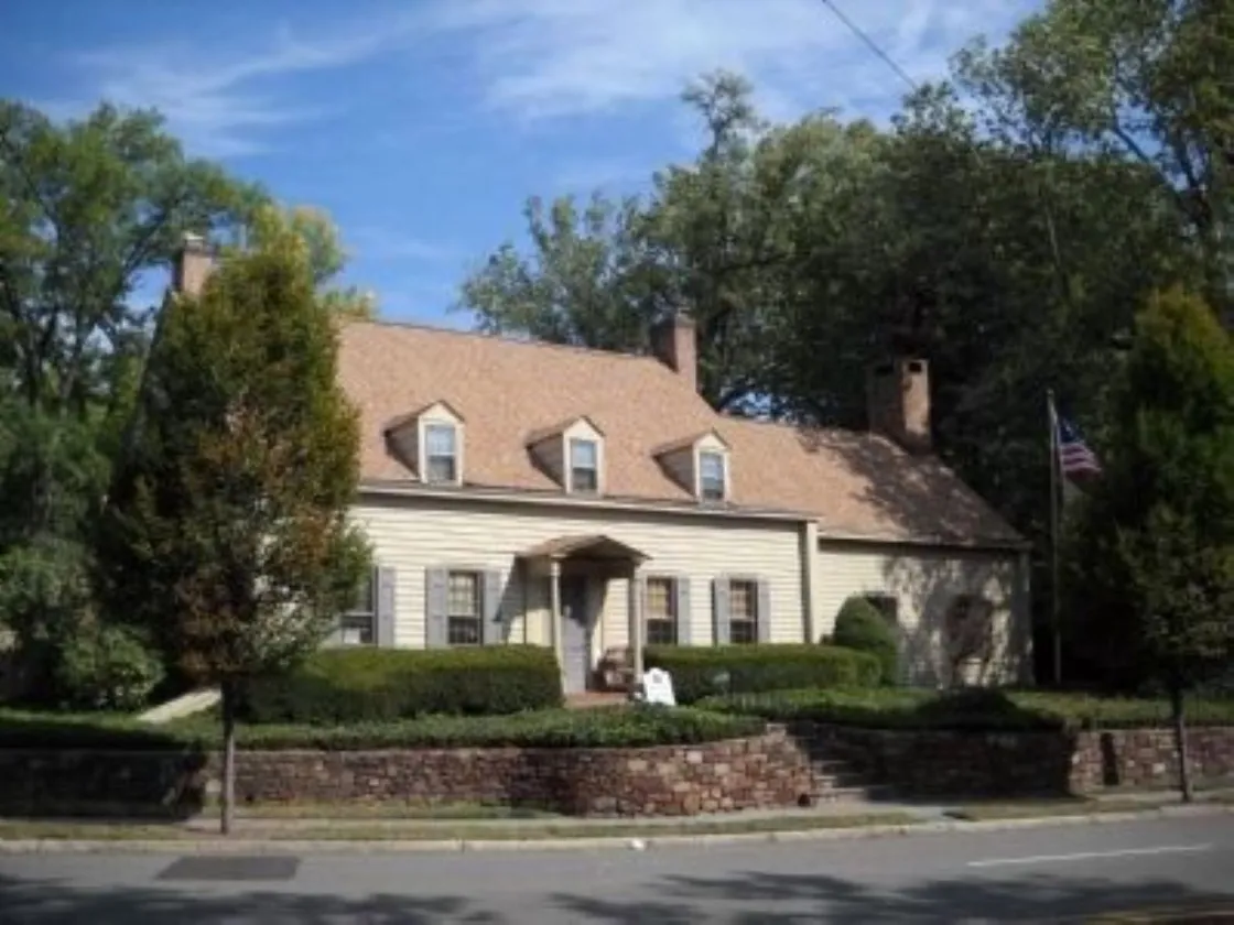 A tan colonial‑style house with dormer windows, brick chimneys, and a columned porch stands behind trimmed hedges and a stone retaining wall, with an American flag flying beside it.