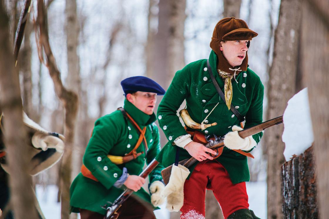 Reenactors in green coats holding rifles in a winter wooded landscape.