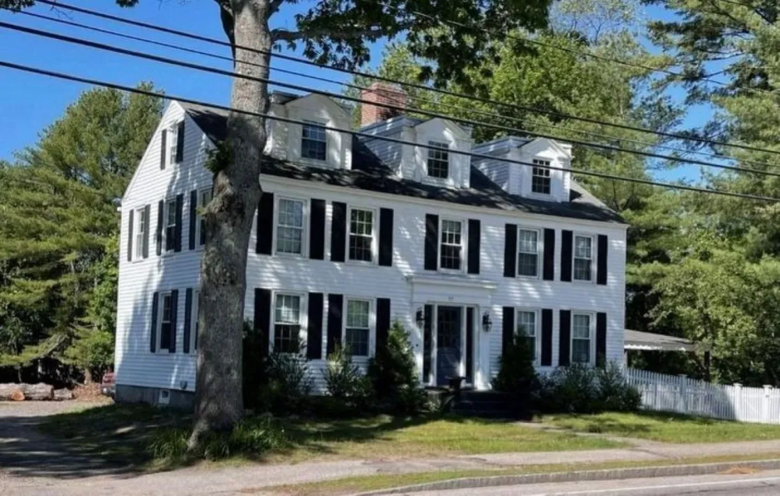 A large white colonial‑style house with black shutters and three dormer windows stands behind a picket fence, surrounded by trees along a roadside.