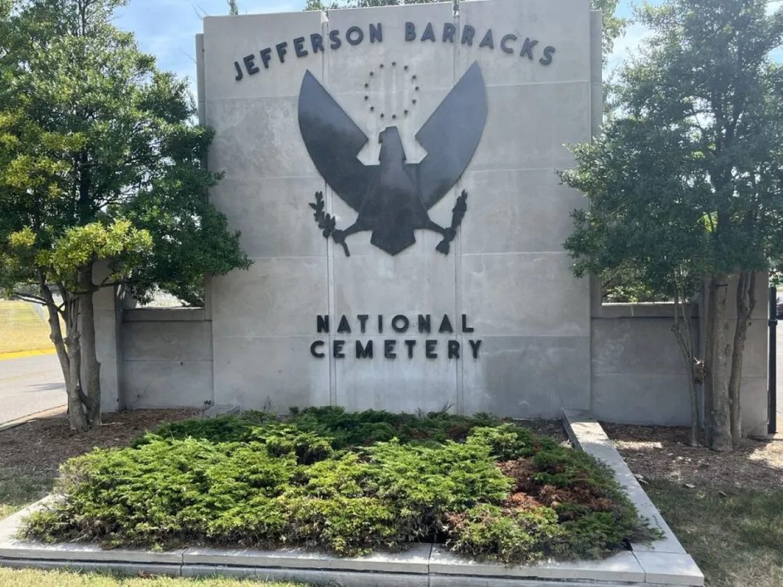 A stone entrance sign for Jefferson Barracks National Cemetery stands among neatly trimmed shrubs and trees. The sign features a carved emblem of an eagle with outstretched wings holding an olive branch and arrows, with stars above its head.