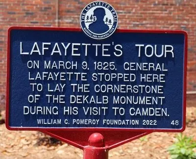 A blue historical marker titled “Lafayette’s Tour” stands in front of a brick wall and plants, describing General Lafayette’s 1825 visit to lay the cornerstone of the DeKalb Monument in Camden.