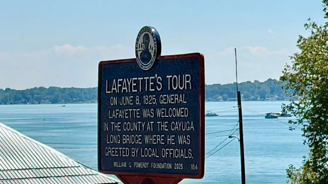 A blue historical marker outlined in red with white lettering titled “Lafayette’s Tour” stands on a lawn in front of a lake commemorating Lafayette's visit to this city.