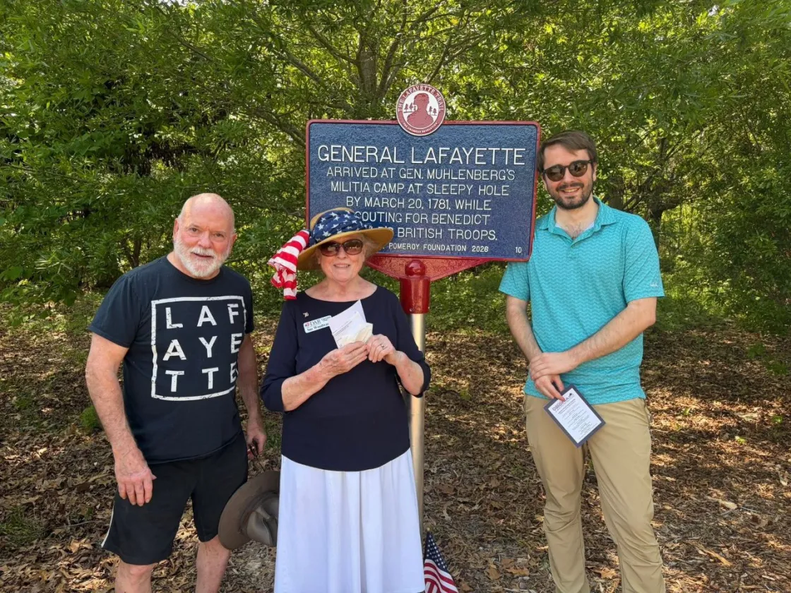 A blue historical marker outlined in red with white lettering titled “General Lafayette” stands on a lawn in front of trees and 3 people commemorating Lafayette's encampment in this city.