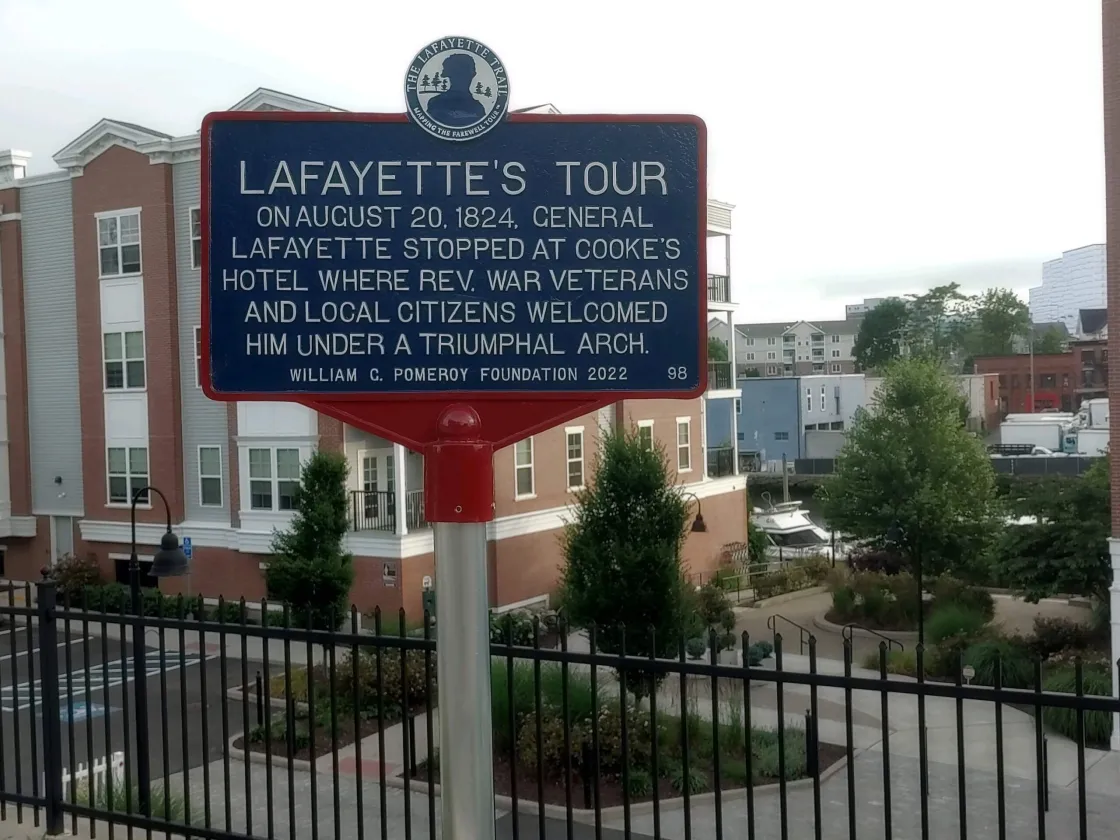 A blue historical marker on a red post describes General Lafayette’s 1824 visit, standing in front of modern brick and white buildings with a fenced walkway and landscaped greenery.