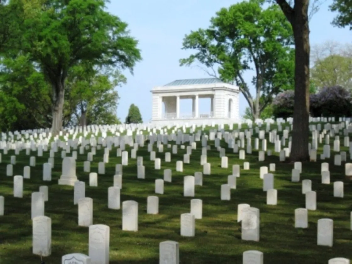 Rows of white headstones line a grassy hillside in a quiet cemetery. Behind them stands a white stone memorial building with columns and a metal roof, surrounded by tall green trees that create a calm, shaded atmosphere.
