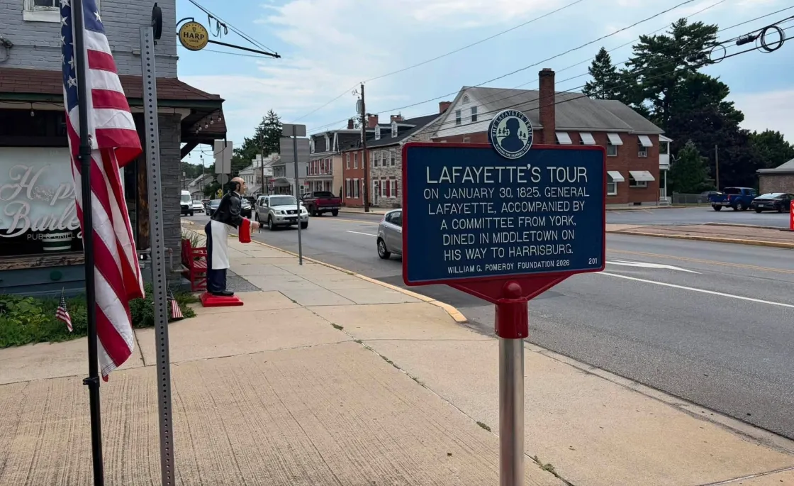 Close up of a sign that contains a description of Lafayette's Tour with a street lined with buildings behind it.