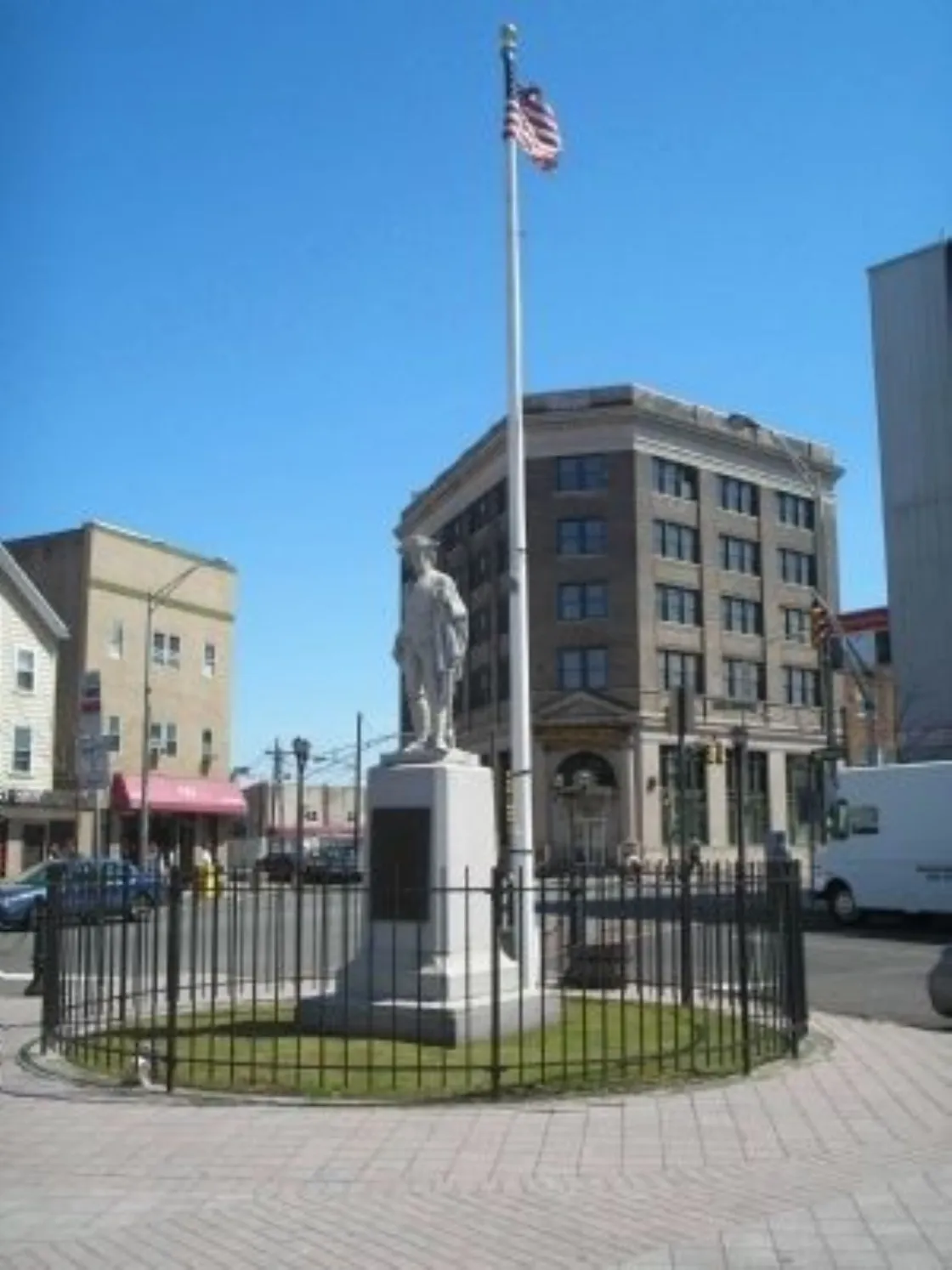A colonial era militia statue enclosed by a circular black fence stands in a small plaza with an American flag behind it, surrounded by multi‑story city buildings under a bright blue sky.