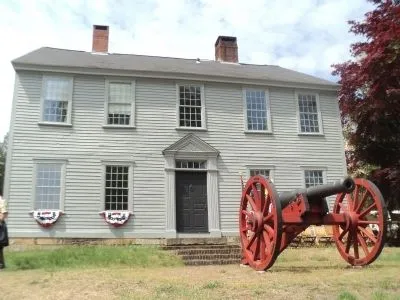 Colonial era 2-story manor house with 9 windows trimmed in white, with 2 chimneys and a red cannon in the front yard.