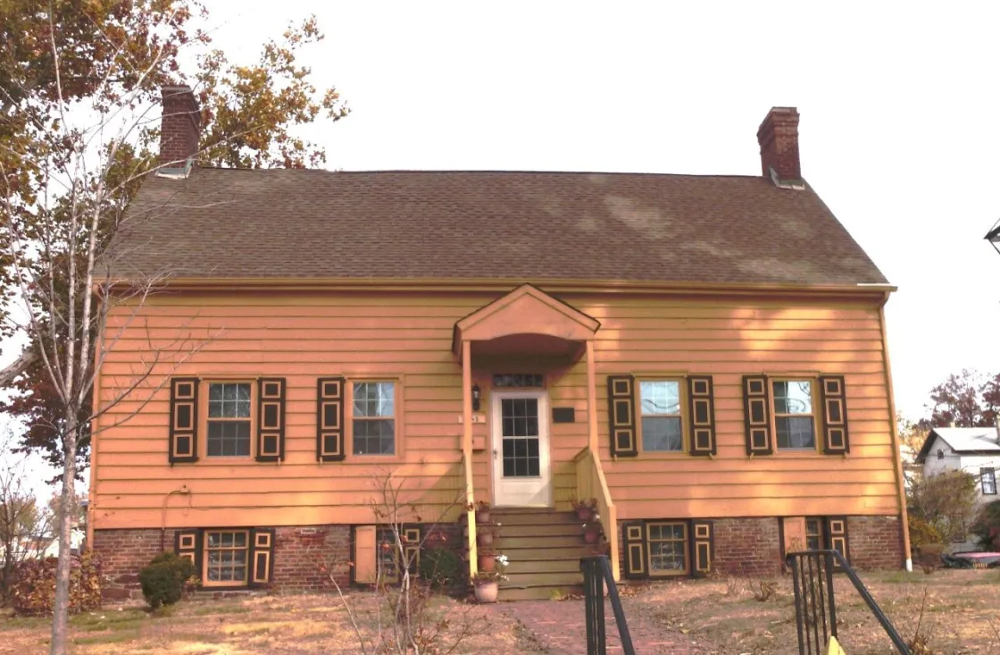 A two‑story yellow colonial house with dark brown trim and twin brick chimneys stands behind a small front porch and brick walkway.