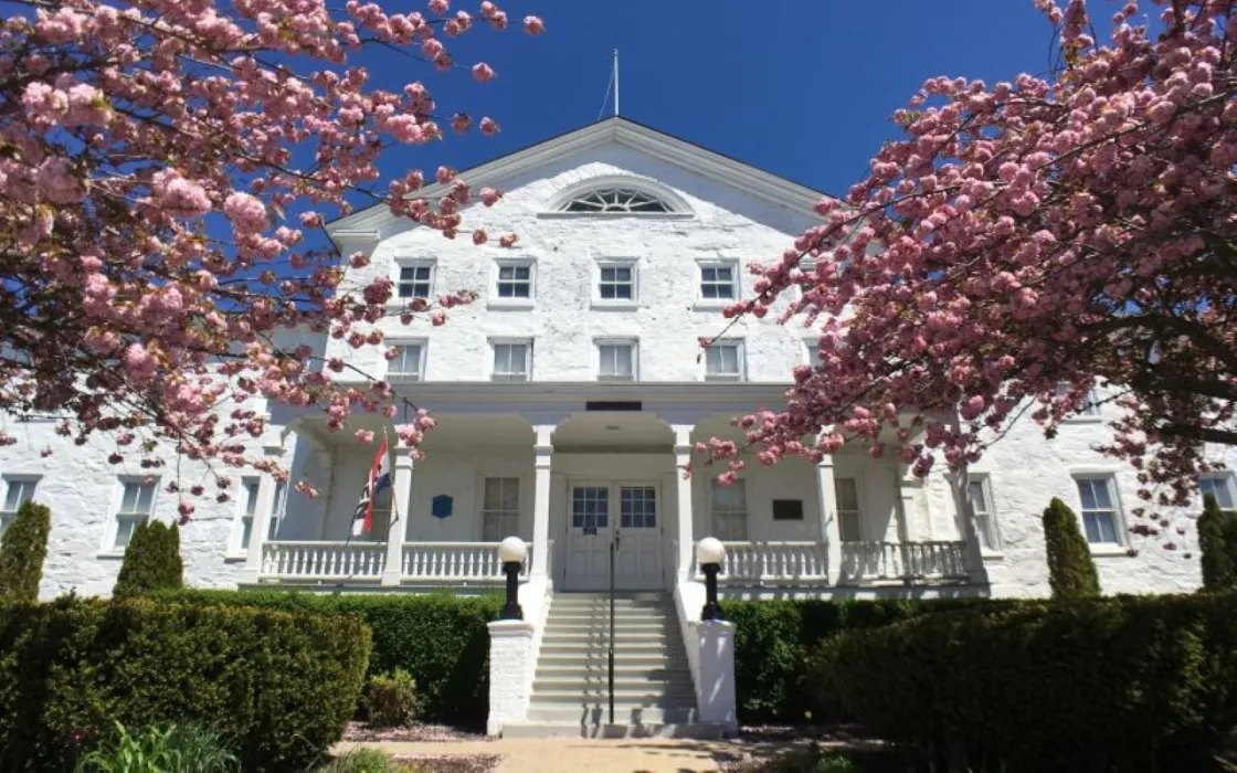 A white colonial‑style building with a central staircase and double doors is framed by bright pink flowering trees, with an American flag near the entrance and a clear blue sky overhead.