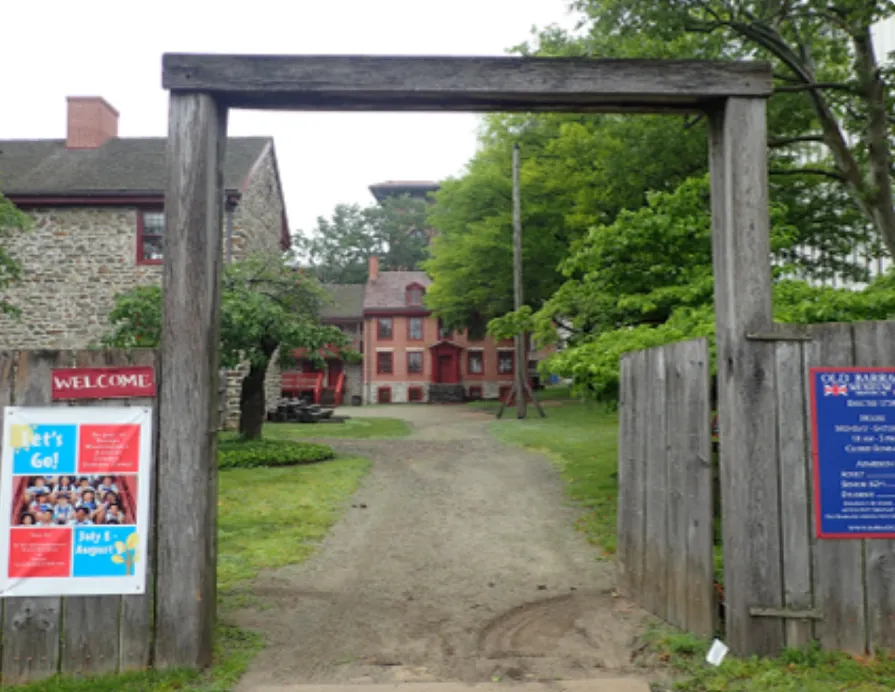 A wooden entrance gate stands at the Old Barracks Museum, built from two tall posts and a horizontal beam.  Beyond the gate, a gravel path leads to historic stone and brick buildings with red trim, surrounded by trees and greenery.