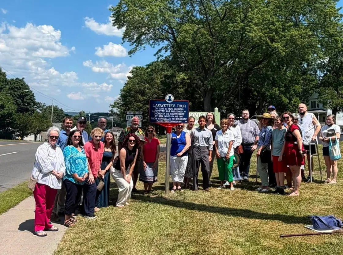 A blue historical marker outlined in red with white lettering titled “Lafayette’s Tour” stands on a lawn in front of large tree with a group of peoople commemorating Lafayette's visit to this city.