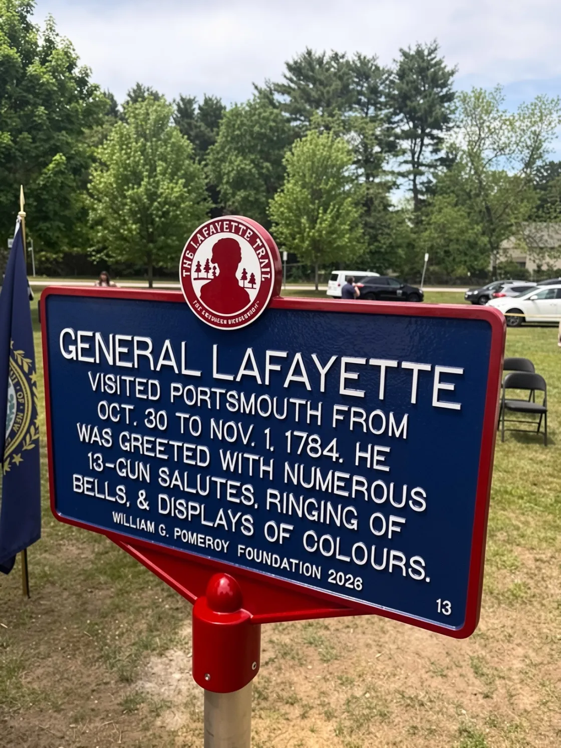 A blue historical marker on a red post describes General Lafayette’s 1824 visit, standing in front of modern brick and white buildings with a fenced walkway and landscaped greenery.
