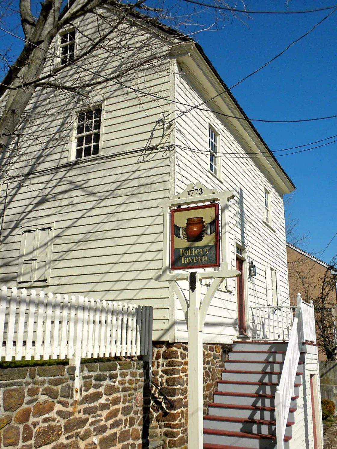 A white historic salt box style building with a stone foundation and an exterior stairway stands behind a sign reading “Potter’s Tavern 1773” topped with a brown pot illustration.