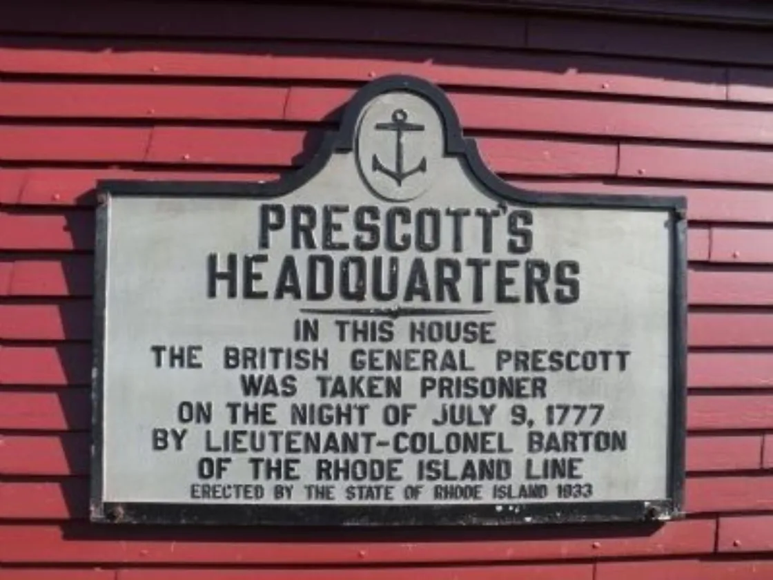 A rectangular historical marker to British General Prescott mounted on a red wooden wall.