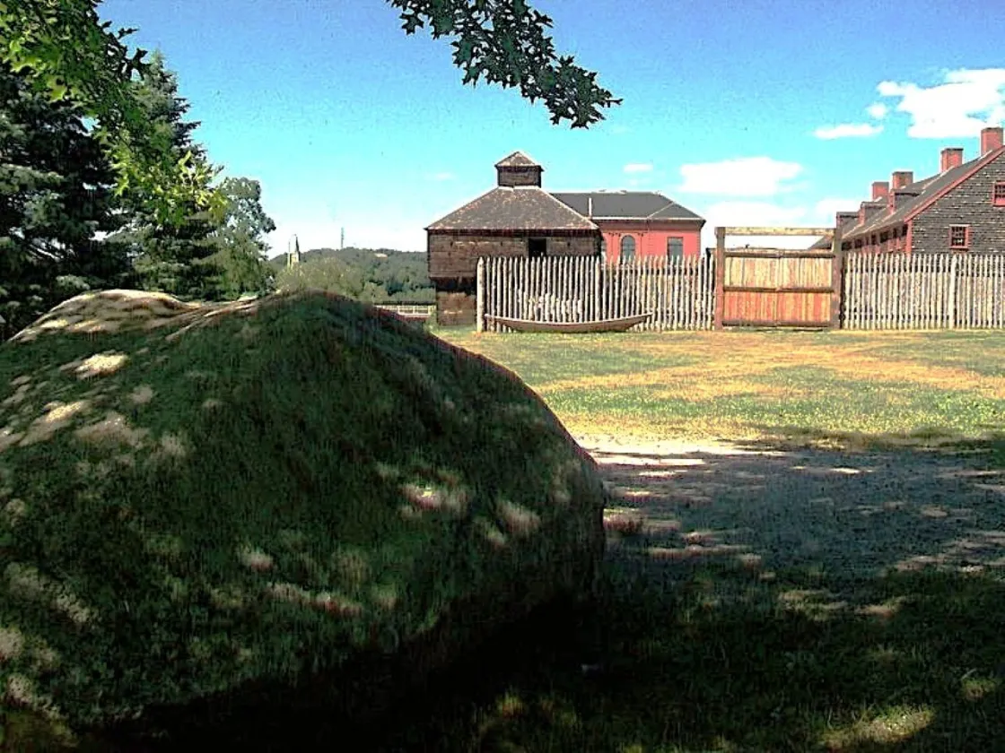 A large boulder sits in the shade before a reconstructed colonial-era fort enclosed by a wooden palisade fence, with historic buildings and open grounds under a bright blue sky.