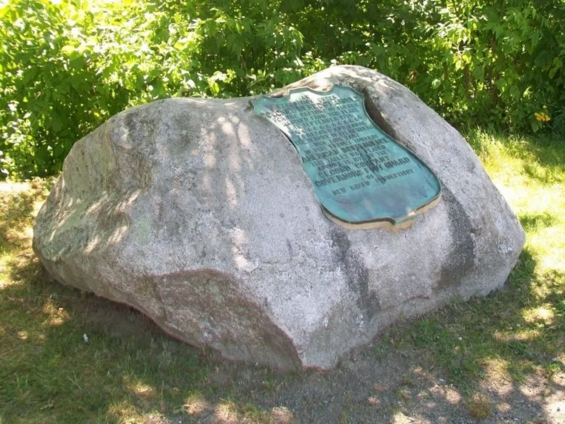 A large boulder with bronze plaque sits on open grounds under a bright blue sky.