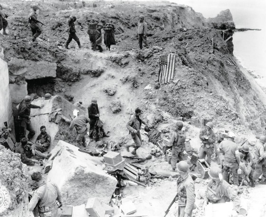 Black and white photo of group of solider and captives atop cliffs with U.S. flag