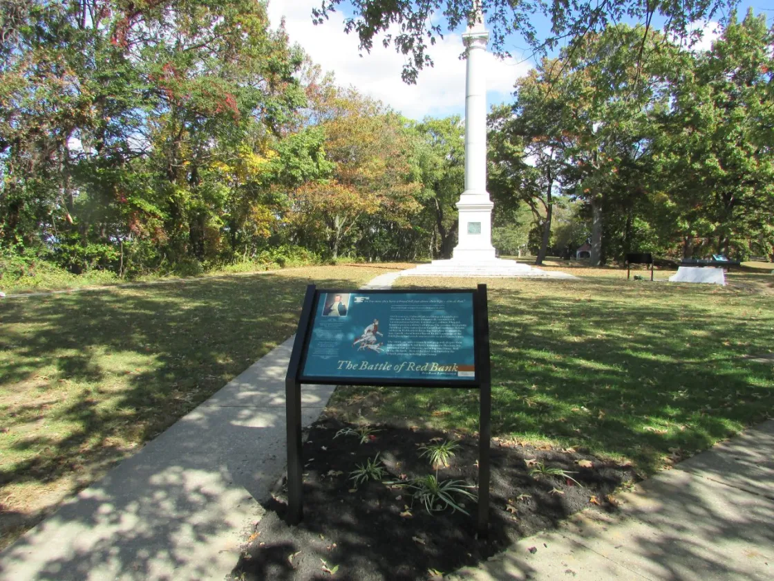 A tall white monument stands behind an informational sign about the Battle of Red Bank in a grassy area with autumn trees.