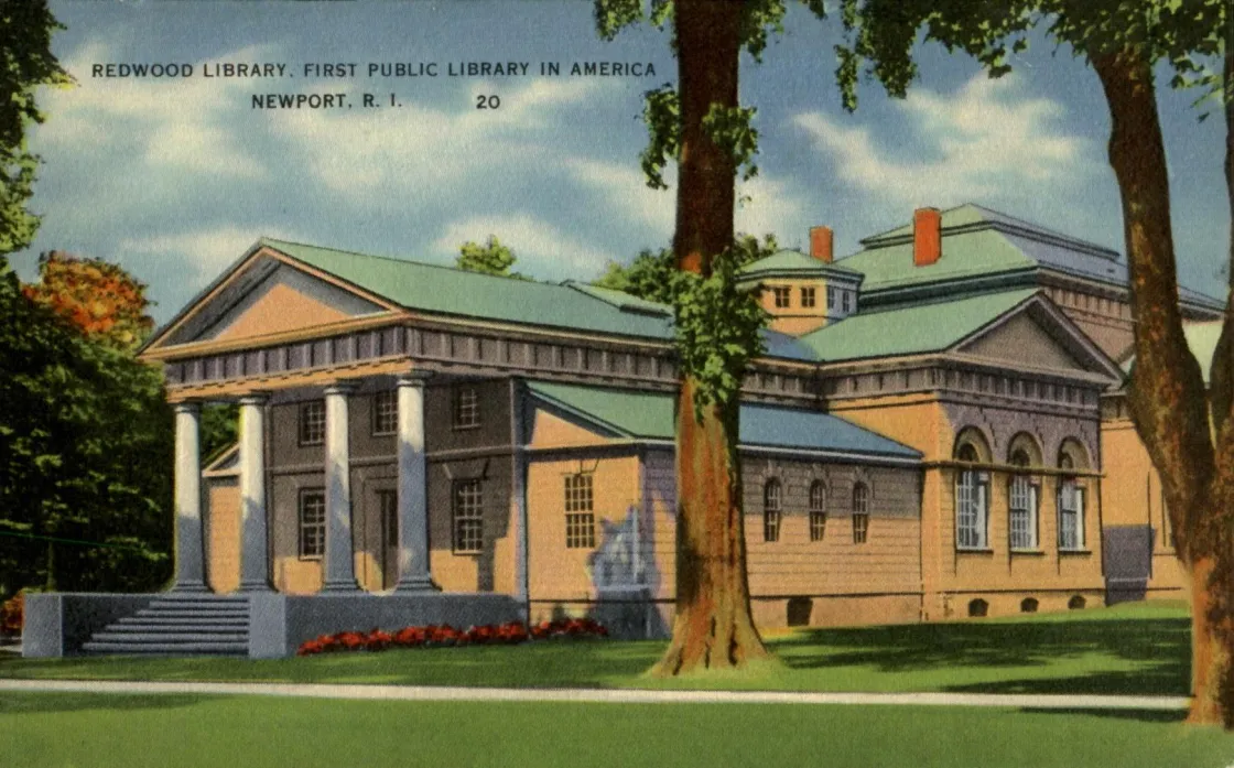 A classical brick building with a columned portico and triangular pediment stands behind a small lamp post, surrounded by trees and greenery.