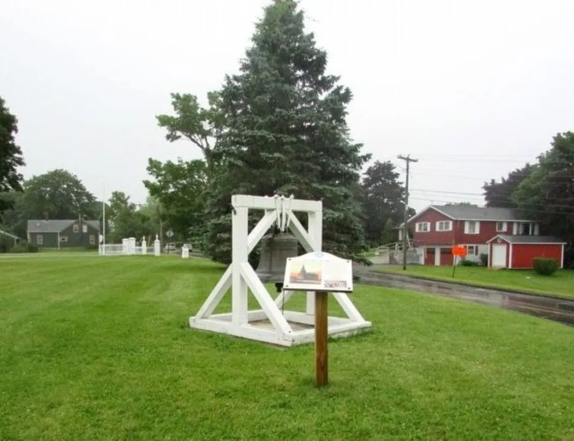 A historic bell hangs in a white wooden frame on a grassy village green, with a large evergreen tree behind it and red houses lining a quiet road.