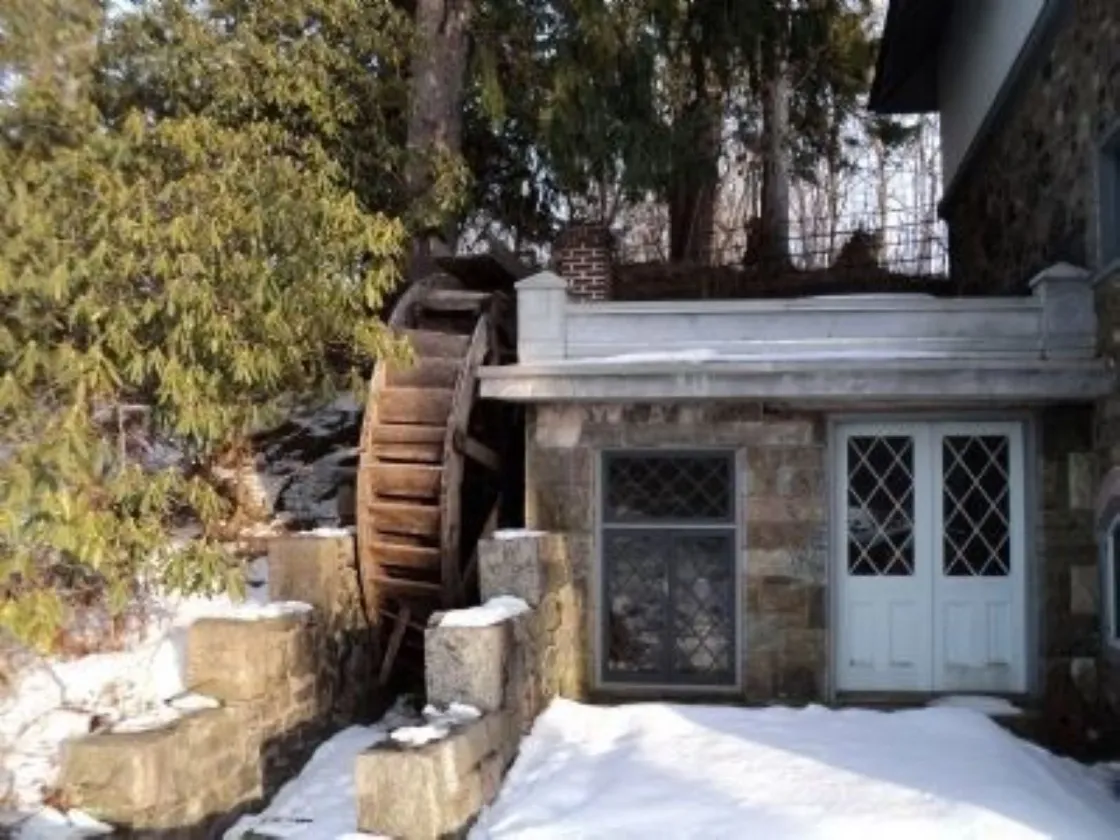 A rustic stone mill with a large wooden water wheel stands in the snow among evergreen trees.