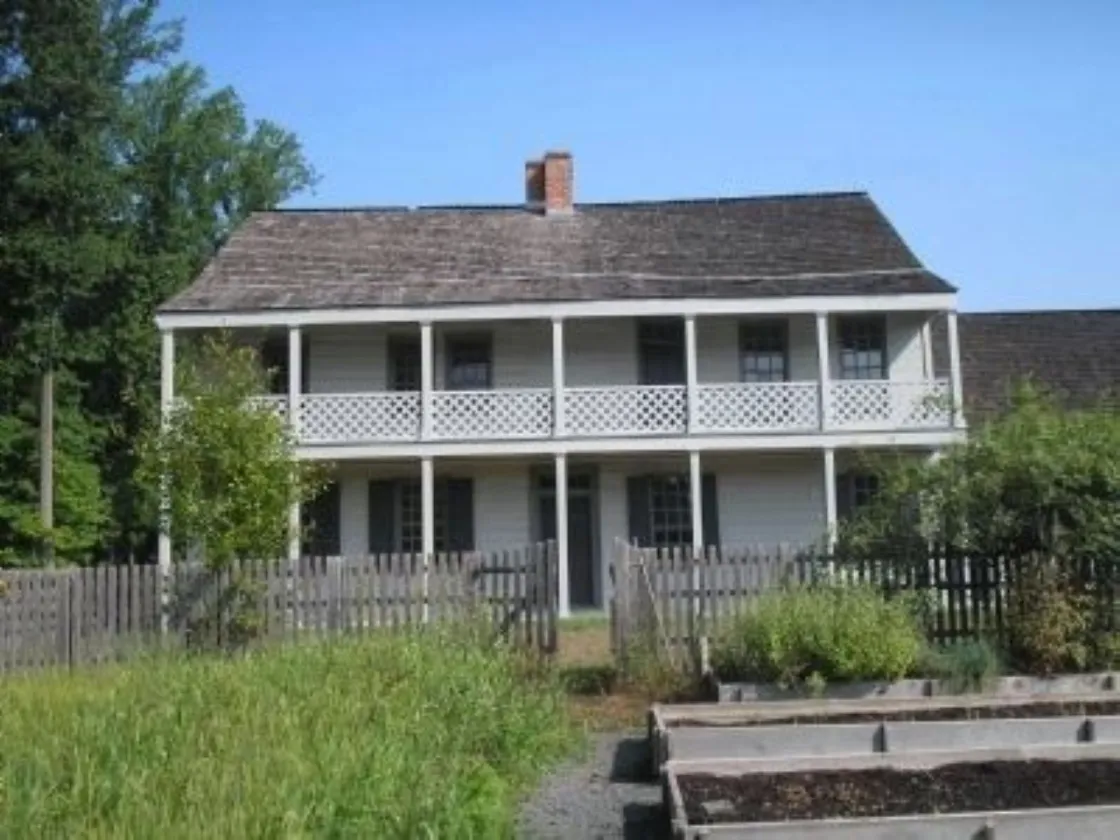 A two‑story wooden house with a long lattice‑railed balcony stands behind a fenced yard with raised garden beds and surrounding trees.