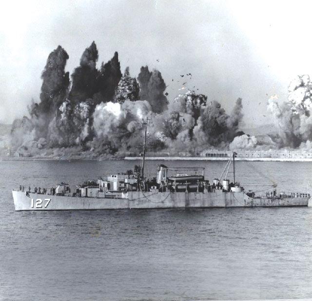 Black and white photo shows a large explosion in the background behind a ship with the number 127.