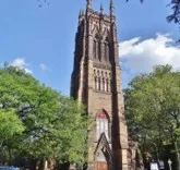 A tall brown stone Gothic‑style church tower with pointed arches and ornate spires rises above trees under a bright blue sky.