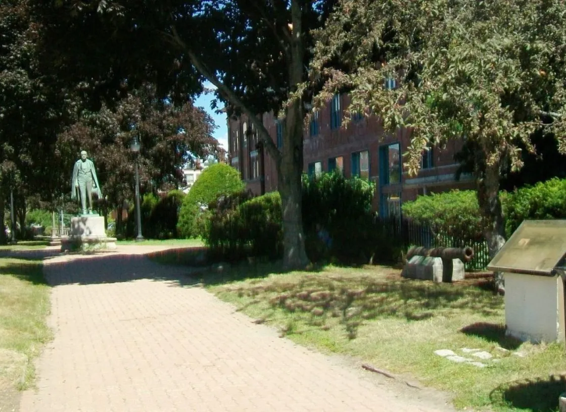 A brick pathway shaded by mature trees leads through a historic park featuring a bronze statue, an interpretive marker, and a small cannon displayed beside a red-brick building on a sunny day.