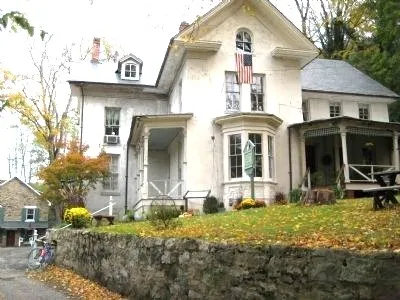 A large light‑colored house with multiple gables, a stone wall in front, and an American flag above the porch sits among autumn trees with fallen leaves.