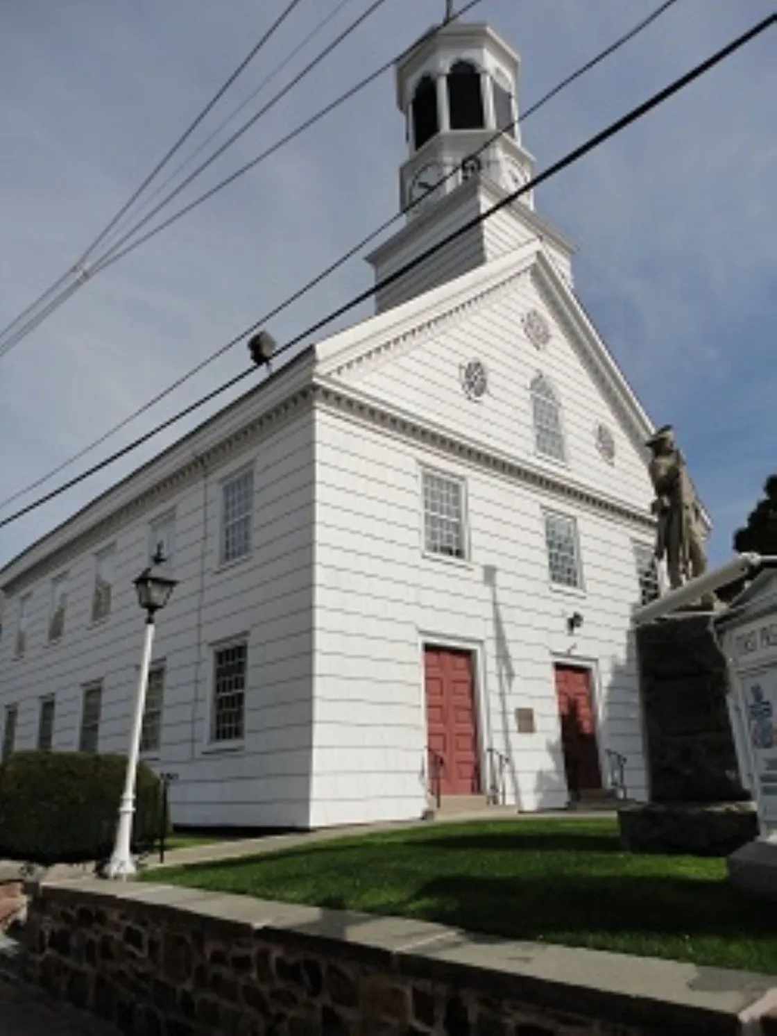 A white wooden church with a tall clock‑tower steeple and red doors stands behind a stone‑bordered lawn with a lamppost and statue of a Revolutionary war soldier.