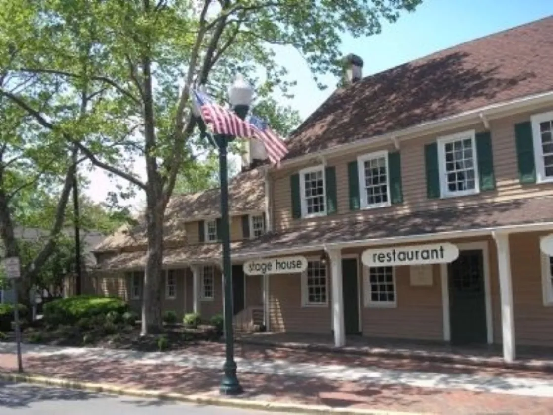 A tan historic building with green shutters and a columned porch stands along a brick sidewalk, shaded by large trees and marked by a streetlamp flying an American flag.