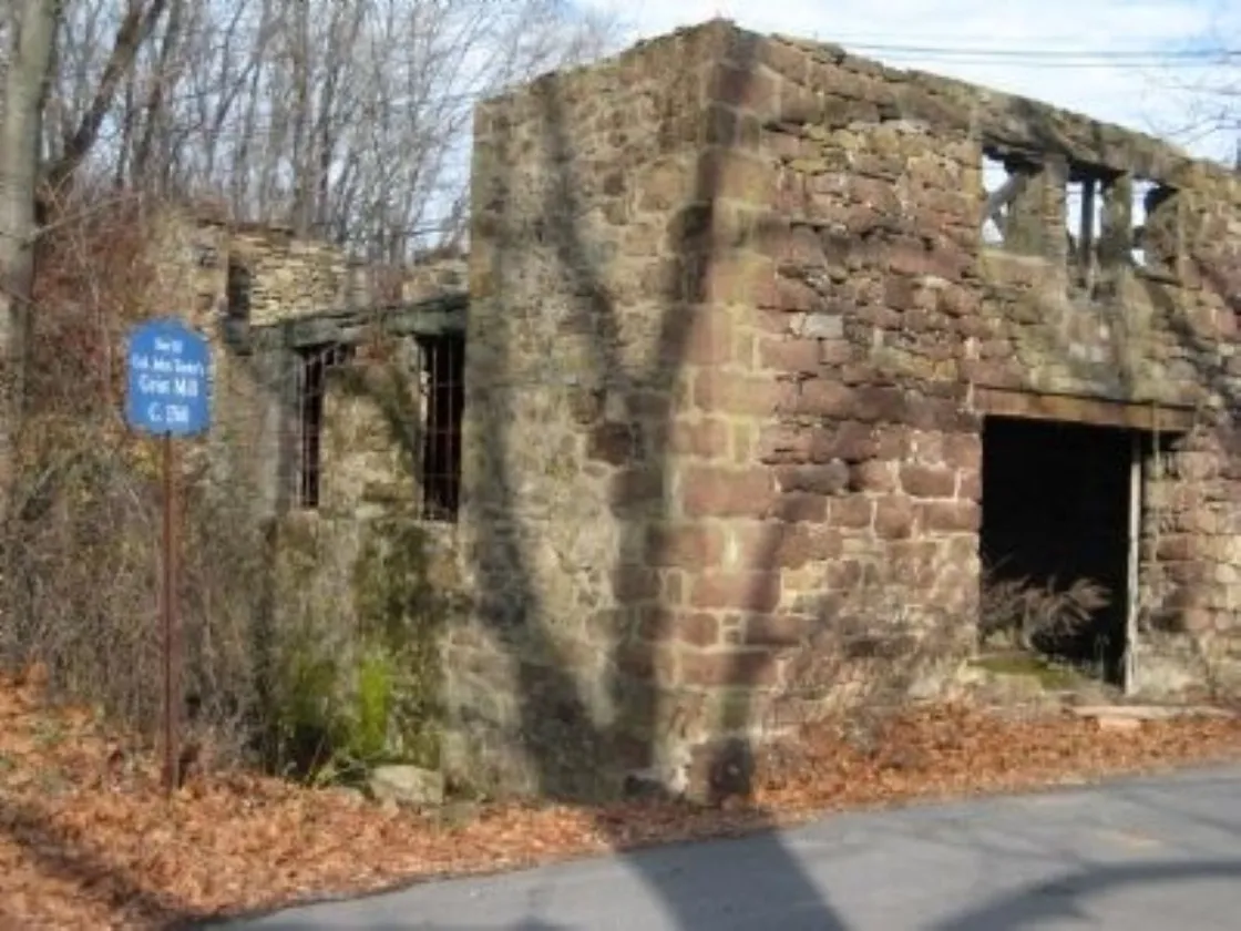 The stone ruins of an old grist mill stand among leafless trees, with a blue historical marker identifying the site beside the weathered walls.