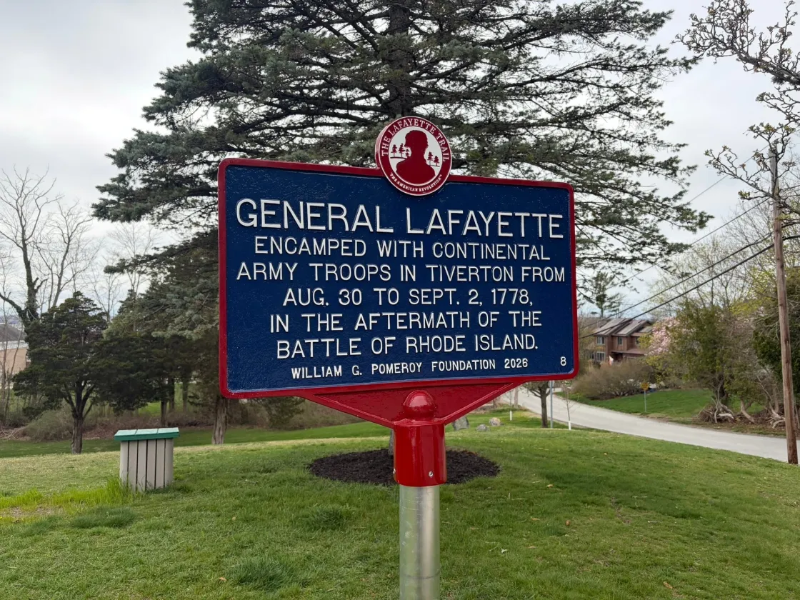 A blue historical marker outlined in red with white lettering titled “General Lafayette” stands on a lawn in front of a tree commemorating Lafayette's encampment in this city.