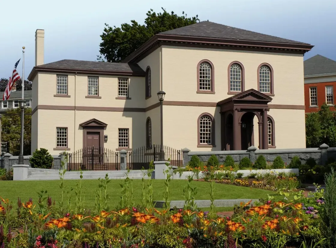 A beige building with brown trim and a columned portico stands behind a garden of bright flowers and neatly trimmed shrubs, bordered by a wrought‑iron fence and stone posts.