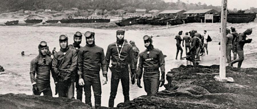 Black and white photo showing group of six men in wet suits standing just off shore, with another group of people in background.