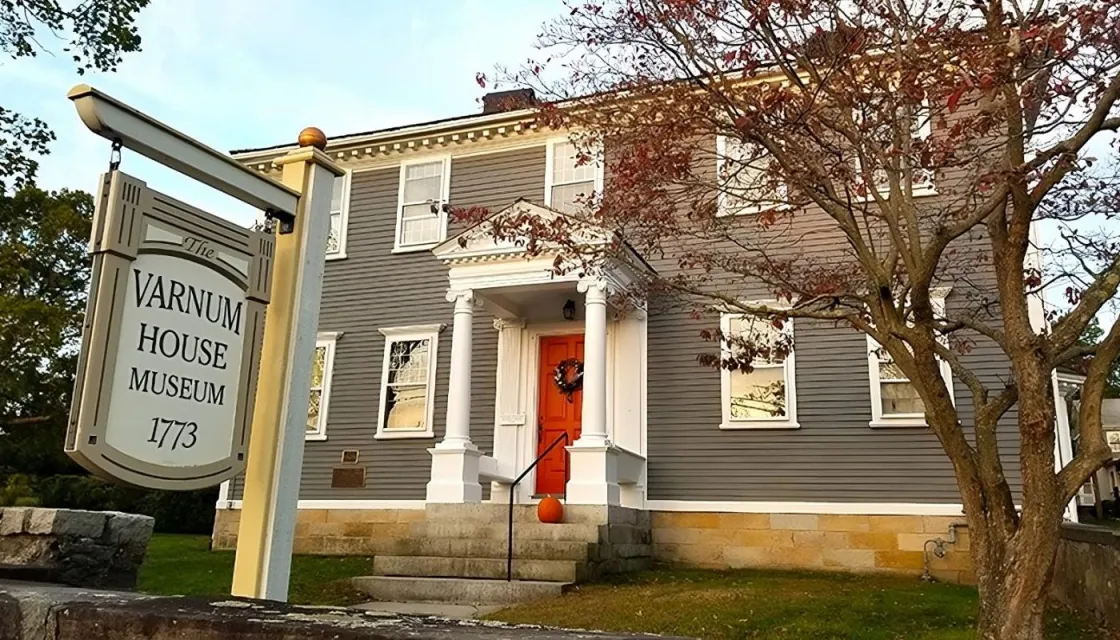 A gray colonial‑era house with white trim and an orange door stands behind stone steps and a small portico. A museum sign out front beneath a tree with sparse autumn leaves.