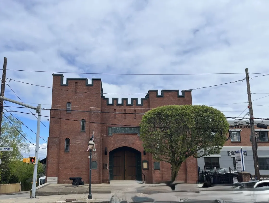 A small fortress like brick building sits on a street corner, with arched wooden doors, battlements, and a nearby cannon. 