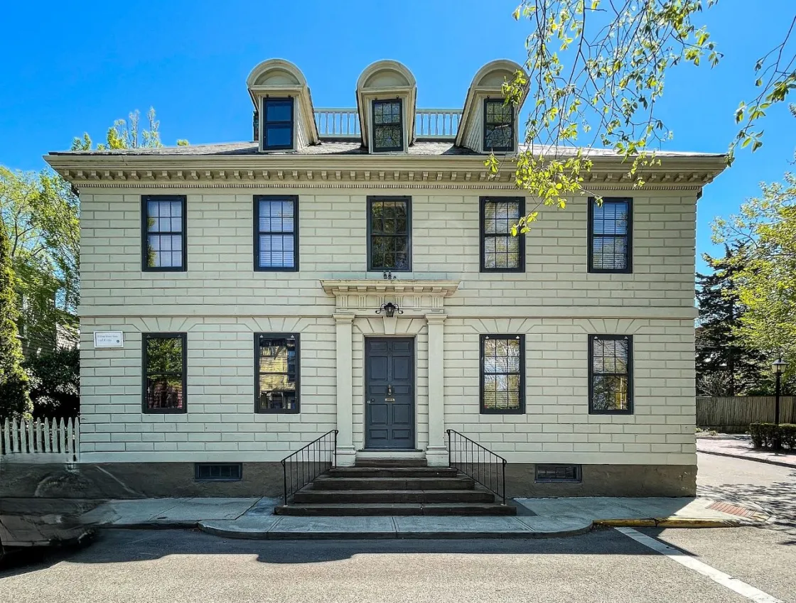 A light‑colored historic building with dark shutters and a central double‑door entrance sits on a corner lot, featuring three dormer windows on the roof and a short staircase leading up from the sidewalk.