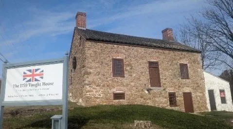 A rectangular stone house with two chimneys stands on a grassy lawn, with a sign in front identifying it as the 1759 Vought House, a Loyalist home from Revolutionary times.