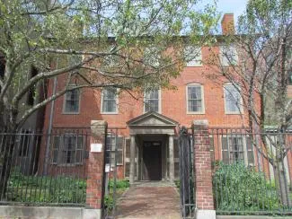 A three‑story, two chimney brick building stands behind a black metal fence and brick pillars, partly shaded by leafy trees.