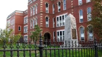 A tall red‑brick historic building with arched windows stands behind a small stone monument shaped like an obelisk. The monument has a metal plaque on its front and is enclosed by a black wrought‑iron fence.