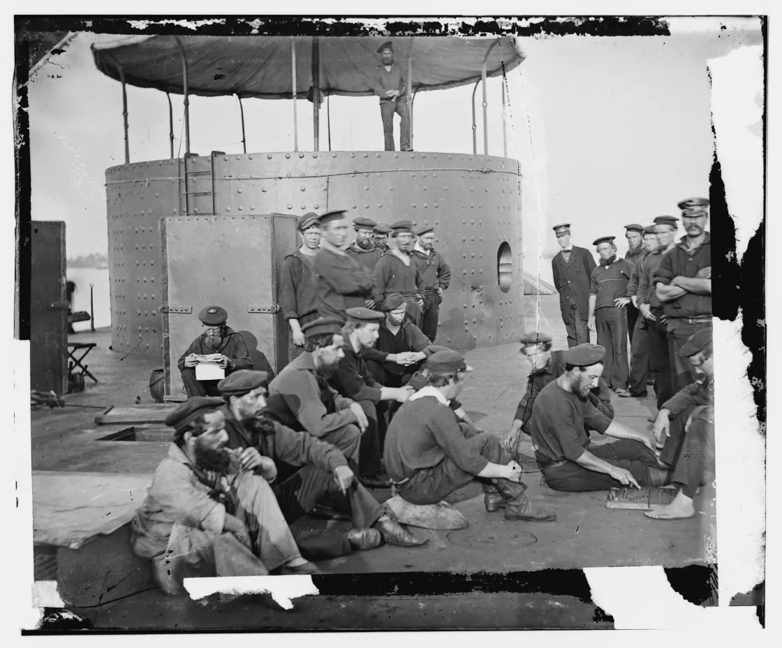 Sailors aboard the USS Monitor relax on the ironclad's deck while anchored in Virginia's James River, some seated and socializing, including crew member Robert Williams standing at the far right.