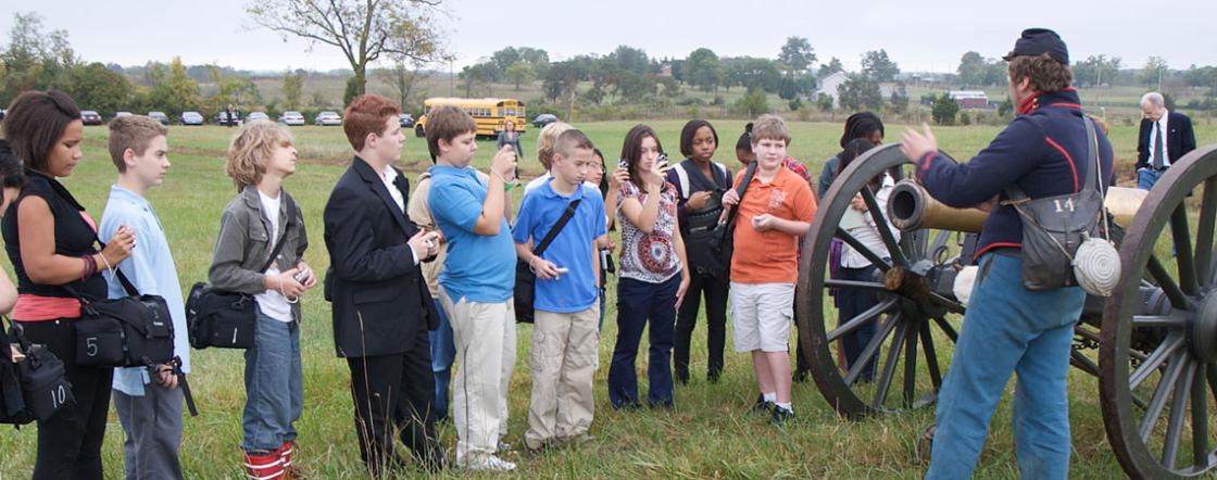 This image depicts a tour guide presenting to a group in front of a battlefield cannon.