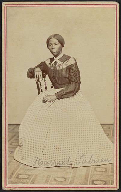 Black woman wearing a dress is seated with her arm along the back of a chair.